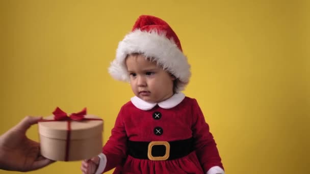 Happy Cheerful Chubby Toddler Baby Girl in Santa Suit Looking On Camera At Yellow Background. Child Play Christmas Scene Celebrating Birthday. Kid open receive surprise gift box present New Year Time — Stock Video