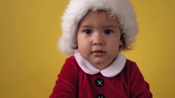 Mignon heureux gai joufflu tout-petit bébé fille en costume de Santa regardant caméra à fond jaune. Scène de jeu d'enfant célébrant l'anniversaire. Kid mord Noël jouets biscuits décoration Nouvel An temps — Video