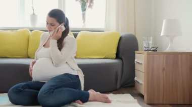 Sevimli hamile kadın, cep telefonuyla doktorla internet üzerinden akıllı telefon görüşmesi yapıyor. Hamilelikteki genç Ledy, evde yerde eğleniyor. Annelik, Sağlık Hizmetleri Konsepti