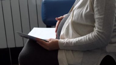 Genç Hamile Koruyucu Maskeli Kadın, hastane için randevu bekleyen koltukta kitap okuyor. Klinikteki jinekoloğu ziyaret et. Muayene Edilmeyi Bekleyen Göbek Sağlığı Kontrolü