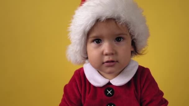 Primer plano emoción lindo feliz alegre regordete bebé niña en traje de Santa mirando en la cámara en el fondo amarillo. Niño jugando a la escena navideña celebrando cumpleaños. El niño se divierte pasar el tiempo de año nuevo — Vídeo de stock