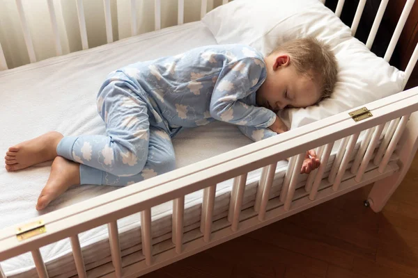 Arriba ver Lindo niño pequeño de 2-3 años de preescolar que duerme dulcemente en una cuna blanca durante el almuerzo, tiempo de descanso en pijama azul con almohada en casa. Infancia, ocio, comodidad, medicina, concepto de salud — Foto de Stock