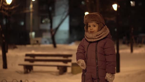 冬、休暇、ゲーム、家族の概念-本格的な幸せな小さな就学前の真ん中の計画幼児の女の子は白い雪を食べる暗い夜の公園で楽しみを持って、寒い霜の天気で雪玉を彫刻 — ストック動画