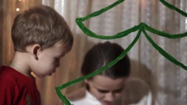 Otantik sevimli bir anne ve iki küçük anaokulu öğrencisi kız ve erkek kardeş 2-4 yaşlarında pencereye Noel ağacı çiziyor. Çocuklu genç bir kadın kar taneleri yapıştırıyor. Xmas, yeni yıl, kış konsepti.