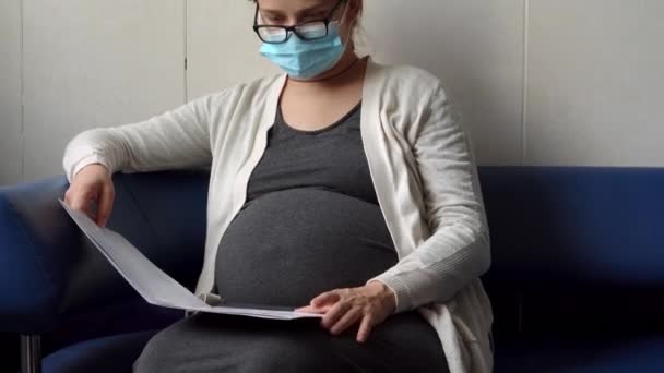 Jeune femme enceinte en médecine masque protecteur lecture sur canapé en attente de rendez-vous contrôle médical sur l'hôpital. Visitez Gynécologue Docteur à la clinique. Examiner le bilan de santé du ventre attendu — Video