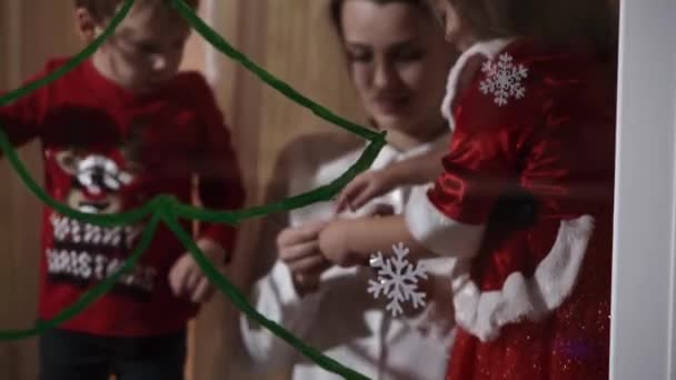 Hiteles aranyos anya és két kis óvodai kiskorú gyerekek testvérek lány és fiú 2-4 éves felhívni karácsonyfa az ablakon. fiatal nő gyerekekkel ragasztó hópelyhek. Karácsony, Szilveszter, téli koncepció. — Stock videók
