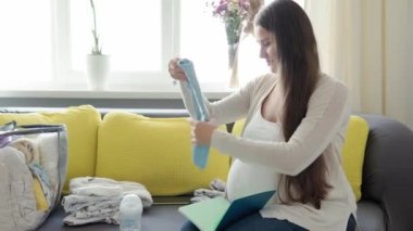 Sevimli Hamile Kadın Doğum Hastanesi için Toplanma Listesi Hazırlık Çantası Yazıyor. Hamilelikteki genç Ledy evde sarı gri koltukta eğleniyor. Annelik, Sağlık Hizmetleri Konsepti