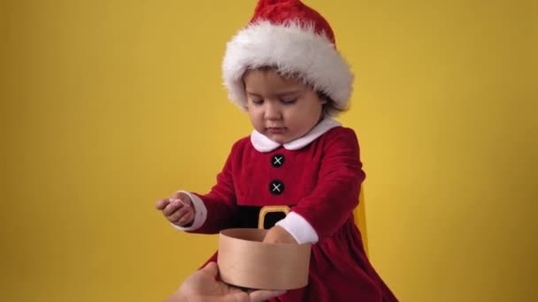 Joyeux joyeux joufflu tout-petit bébé fille en costume Santa regardant la caméra à fond jaune. Jeu d'enfant Scène de Noël Célébration d'anniversaire. Enfant ouvert recevoir surprise boîte cadeau cadeau Nouvel An temps — Video