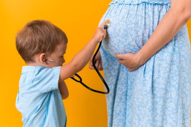 Annelik, sıcak yaz - kesilmiş portre hamile anne tanınamaz kadın mavi elbise küçük oğlan kardeş hamile anne rolü oynuyor steteskop doğmamış anne göbeği sarı arka planda.