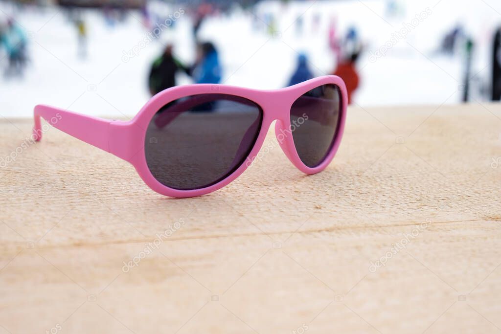 Pink-rimmed sunglasses on wooden slope in apres-ski bar or cafe, with ski slope in background, copy space. Concept of winter sports, leisure, recreation, relaxation in resort.