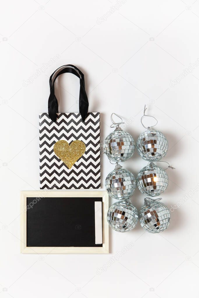 Christmas gift bag with gold heart, six little disco balls, chalk board on white background, copy space. Festive, New Year, sales, shopping concept. Vertical, flat lay. Top view.