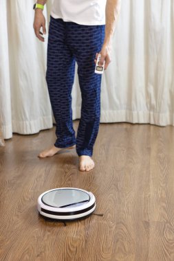 Adam ev temizliği yapıyor, uzaktan kumandayla kablosuz robot elektrik süpürgesini açıyor. Evdeki günlük temizlik kavramı, evdeki erkeklere yardım etmek, ev işleri, hijyen. Yumuşak seçici odak.