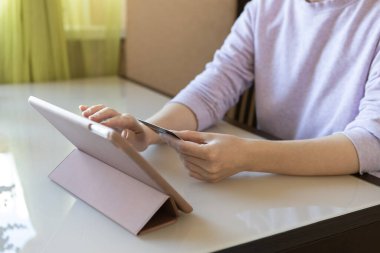 Beyaz, tanınmayan genç bir kadın online mağazadan alışveriş yapıyor, tablet ve kredi kartı kullanıyor, masada oturuyor. Çevrimiçi alışveriş kavramı, evde kalmak, güvende olmak, e-ticaret, teknolojiyi kullanmak.