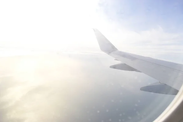 Airplane wing flying above white clouds obscure light from the sun. View from aircraft window.