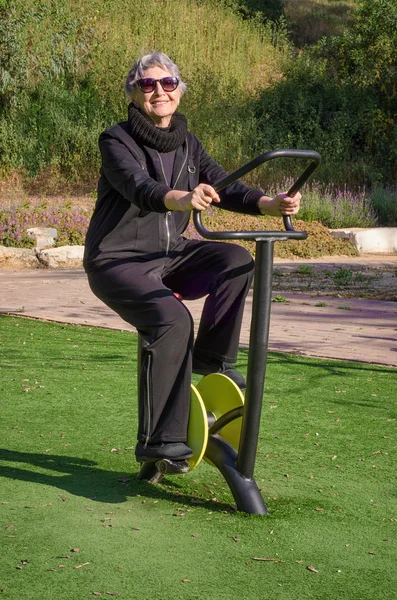 Mulher idosa em terno esporte preto trabalha para fora em treinador ciclo de fiação em seu parque local — Fotografia de Stock