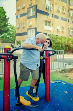 Emekli bir adam, yakınındaki bir parktaki açık hava spor salonunda antrenman yapmaya başlıyor. Egzersizin faydalı olduğunu biliyor, özellikle COVID-19 sırasında. Ücretli bir yardımcı ona yardım eder. İkisi de koruyucu maske takıyor..