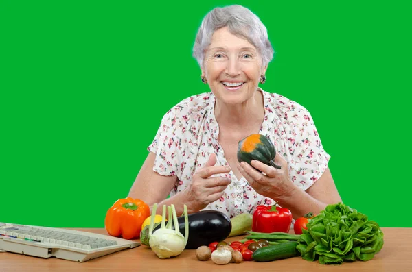 A cheerful elderly vegetarian woman sits at a table with fresh vegetables and looks at the camera. Green background.