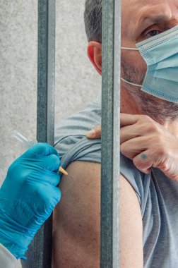 Cezaevlerindeki Sağlık Tedbirleri: Eldivenli El Yöneticileri Barlar Arasında Maskeli Mahkûma Aşı Alıyor, Mahkumları Bulaşıcı Hastalıklardan Koruma Çabaları Gösteriliyor.