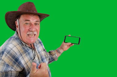 Kahverengi şapkalı deneyimli bir çiftçi yeşil ekranlı akıllı bir telefonu tutarken kendinden emin ve bilgili bir görünüm sergiliyor ve kolayca kandırılamayacağını açıkça ifade ediyor. Yeşil arkaplana göre ayarla.