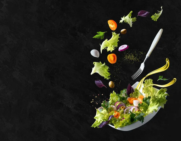 A white plate with salad and floating in the air ingredients: olives, lettuce, onion, tomato, mozzarella cheese, parsley, basil and olive oil. Black background. Copy space.