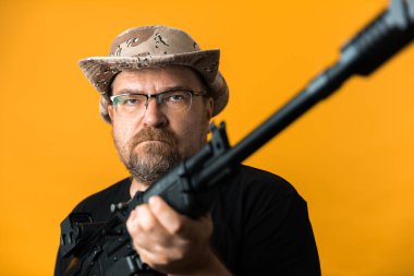 Siyah tişörtlü ve şapkalı orta yaşlı bir adam sarı arka planda..