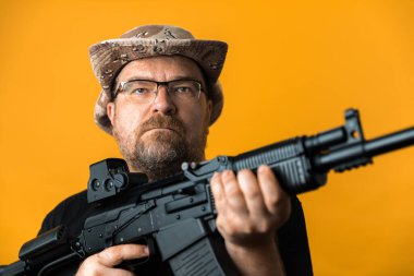 Siyah tişörtlü ve şapkalı orta yaşlı bir adam sarı arka planda..