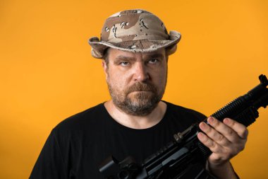 Siyah tişörtlü ve şapkalı orta yaşlı bir adam sarı arka planda..