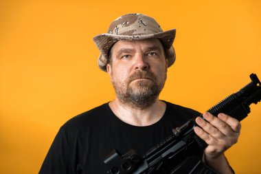 Siyah tişörtlü ve şapkalı orta yaşlı bir adam sarı arka planda..