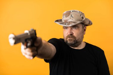 Siyah tişörtlü ve sarı arka planda şapka giyen orta yaşlı bir adam..