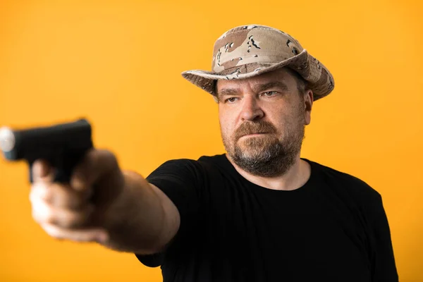 Middle aged man with gun in black t-shirt and hat against yellow background.