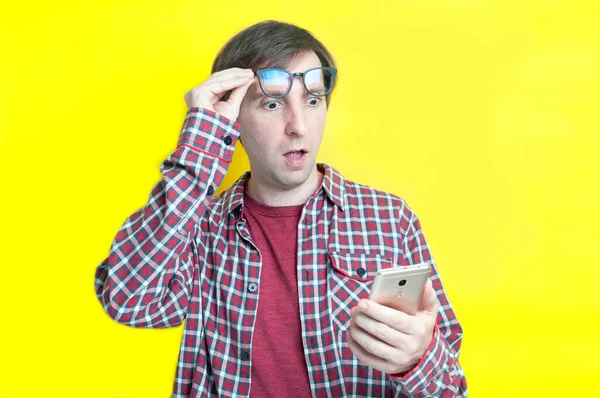 handsome excited man with open mouth in red checkered shirt taking off glasses on forehead and looking at smartphone on yellow background with copy space