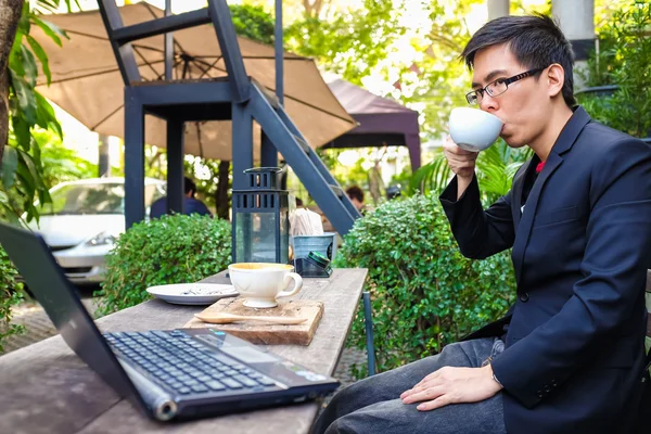 Kafede elinde kahve fincanıyla dizüstü bilgisayarda çalışan bir iş adamı.