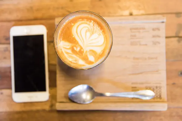 Latte art coffee cup and smartphone on wooden table. — Stock Photo, Image