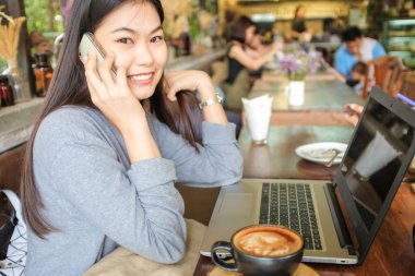 Dizüstü bilgisayar kullanarak ve modern kahve dükkanı iç otururken cep telefonu konuşurken genç mutlu Asyalı kadın