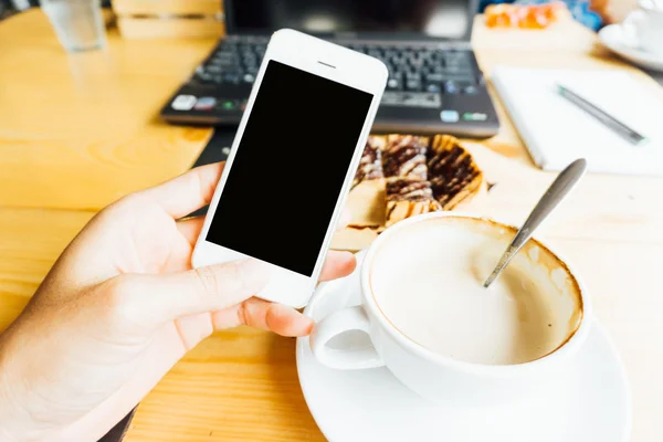 Hand using smartphone in coffee shop with dessert and laptop