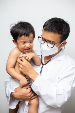 Mutlu Asyalı doktor, beyaz arka planda steteskop muayenesi yapan bebeği muayene ediyor.