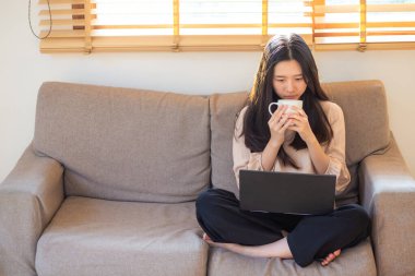 Genç Asyalı kadın rahat bir koltukta otururken bilgisayarla iş bağlantısı kuruyor.