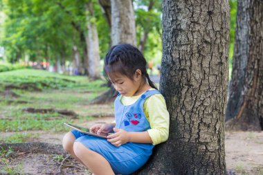 Küçük Asyalı kız ağacın altında oturuyor. Şehir parkında tablet bilgisayar internet tarayıcısı kullanıyor.