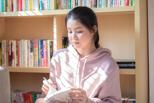 Young asian woman at home sitting in front of book shelf relaxing in her living room reading book, concepts of home and comfort