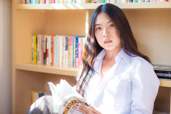 Young asian woman at home sitting in front of book shelf relaxing in her living room reading book, concepts of home and comfort