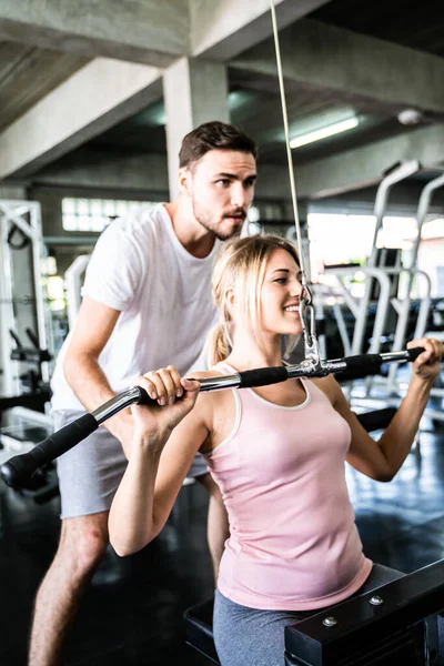 Cheerful young woman wearing pink sports bra while doing chin-up exercise with trainer man in fitness gym