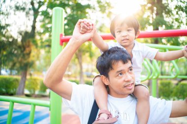 Küçük çocuk dışarıda oyun parkında eğlenirken babasının boynuna biniyor.
