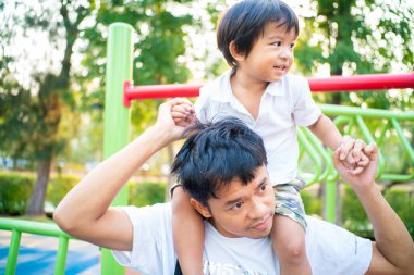 Küçük çocuk dışarıda oyun parkında eğlenirken babasının boynuna biniyor.