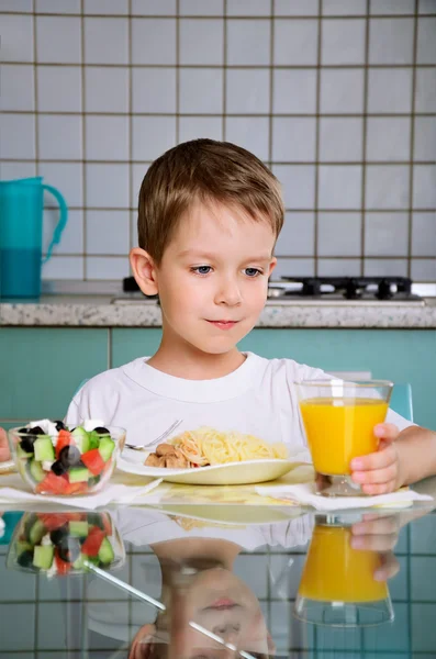 Smiling boy having dinner at the table and takes a glass of oran — Stock Photo, Image