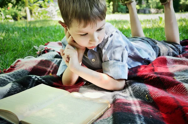 Little boy reading a book while lying on a mat in the park close — Stock Photo, Image