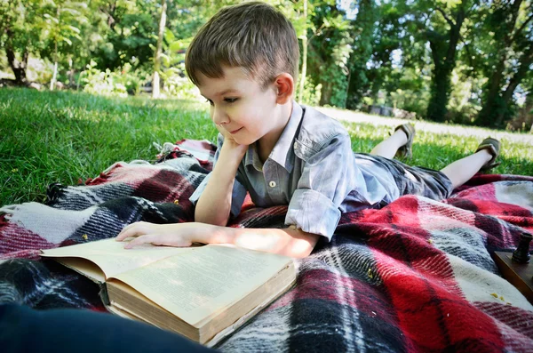 Niño sonriente leyendo un libro mientras está acostado en una alfombra en el parque — Foto de Stock