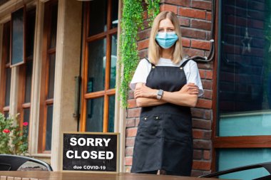 Tıbbi maske takan Kafkasyalı garson kadın ve Üzgünüz kapalıydı. Koronavirüs salgını. Hükümetin restoranları, alışveriş merkezlerini kapatması, temel olmayan hizmetler