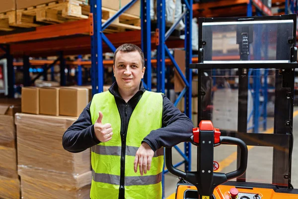 Storehouse employee in uniform working on forklift in modern automatic warehouse. Worker showing thumbs up. Boxes on the shelves of the warehouse. Warehousing, machinery concept. Logistics in stock. High quality photo