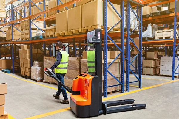 Storehouse man employee in uniform using forklift with box in modern automatic warehouse. Boxes are on the shelves of the warehouse. Warehousing, machinery concept. Logistics in stock.