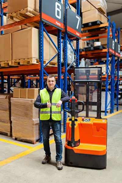 Storehouse employee in uniform working on forklift in modern automatic warehouse. Worker showing thumbs up. Boxes on the shelves of the warehouse. Warehousing, machinery concept. Logistics in stock. High quality photo